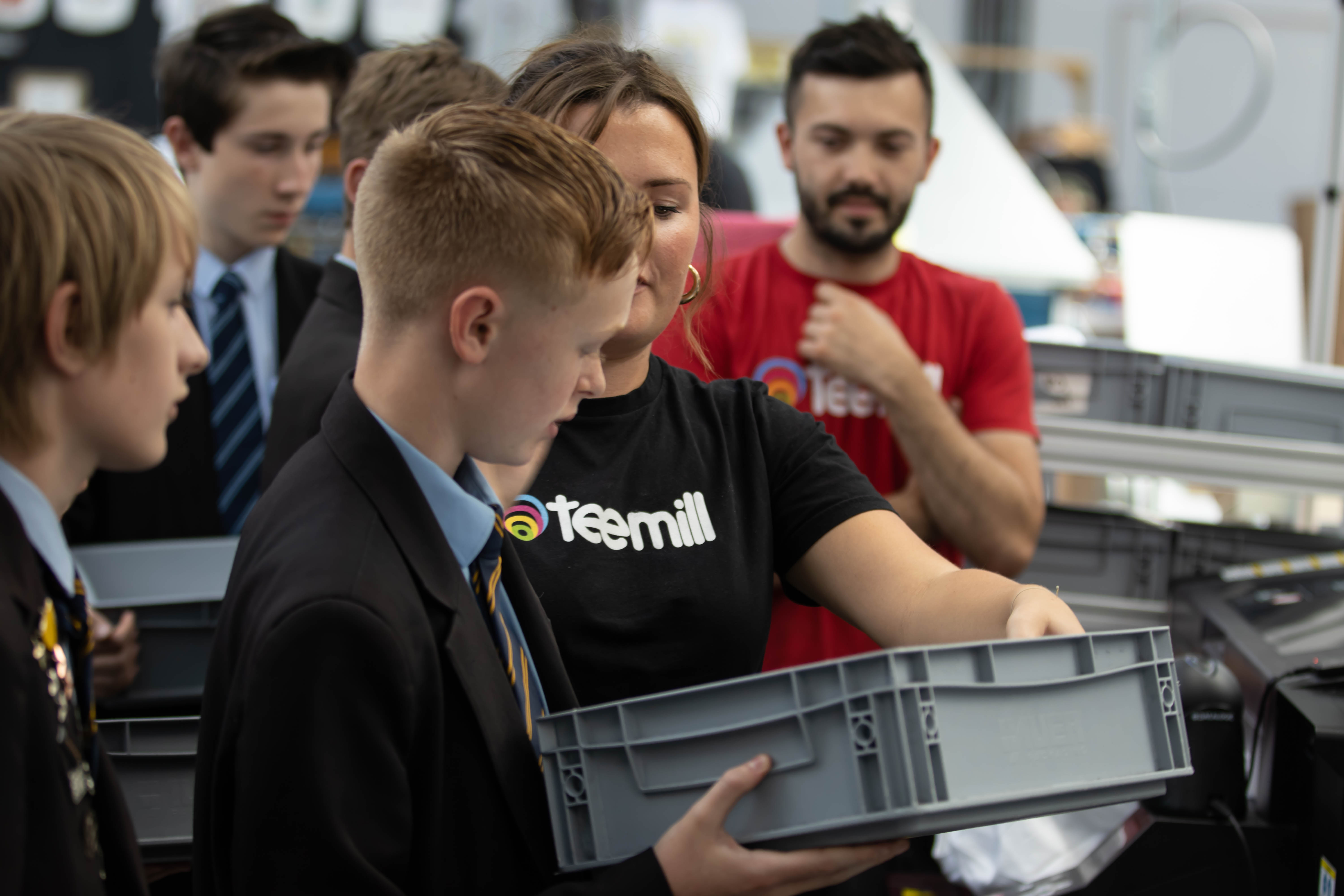 Photo of Teemill staff and pupils in the Teemill factory. A Teemill operative is holding a grey crate, showing it to one of the pupils.